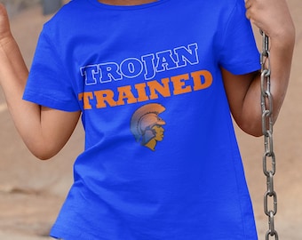 Può includere: Una maglietta blu reale con le parole "TROJAN TRAINED" in lettere bianche e arancioni. Sotto il testo c'è un'illustrazione dorata e marrone della testa di un guerriero troiano. La maglietta è a maniche corte.
