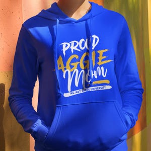 May include: Royal blue hoodie with a front pocket and drawstring. The hoodie features the text "PROUD AGGIE Mom" in white and yellow lettering, with "NC A&T STATE UNIVERSITY" printed below. A casual, comfortable apparel item.