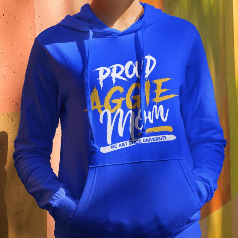 May include: Royal blue hoodie with a front pocket and drawstring. The hoodie features the text "PROUD AGGIE Mom" in white and yellow lettering, with "NC A&T STATE UNIVERSITY" printed below. A casual, comfortable apparel item.