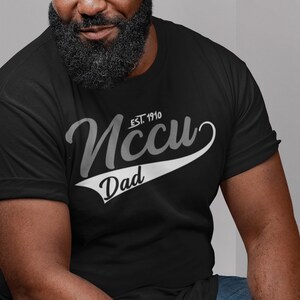 May include: Black t-shirt with white text that reads "NCCU EST. 1910 Dad" in a vintage baseball style font.
