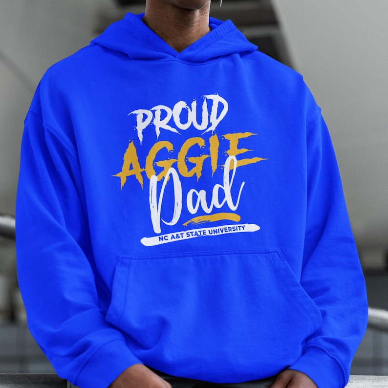 May include: A royal blue hoodie with the words "PROUD AGGIE Dad" in white and gold lettering. The text "NC A&T STATE UNIVERSITY" is printed below the word "Dad". The hoodie has a front pocket and a hood.