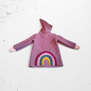 Könnte beinhalten: Ein mauvefarbener Kapuzenmantel mit langen Ärmeln und einem bunten Regenbogen-Applikation. Die Ärmel haben rosa Bündchen. Der Mantel ist für Kinder konzipiert und wird auf weißem Hintergrund präsentiert.