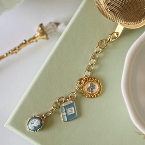 May include: A gold-toned metal tea infuser featuring a chain attachment adorned with three charms: a small blue and white oval cameo, a light blue book with white pages, and a round gold-rimmed charm with a green floral design.