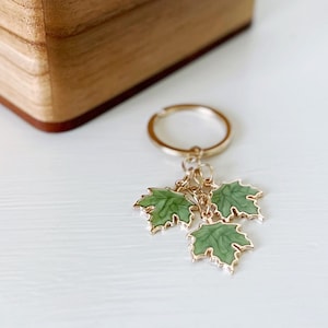 May include: A gold key chain with three green enamel maple leaf charms. The keychain is on a white surface.