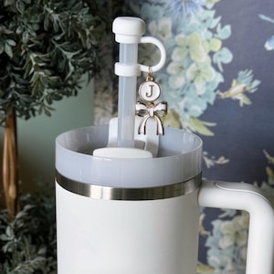 May include: White travel mug with a stainless steel band around the middle. The mug has a white lid with a clear plastic straw and a white handle. A white charm with a gold bow and a gold circle with the letter "J" is attached to the straw.