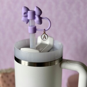 May include: A white travel mug with a stainless steel band around the top. The mug has a clear lid with a purple bow and a straw. The straw has a charm with the letter "A" on it.