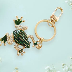 May include: A gold-tone keychain featuring a green frog with rhinestones. The frog is sitting on a gold-tone ring with a lobster clasp.