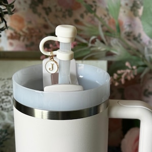 May include: A white insulated tumbler with a light blue rim and handle. The lid features a white straw and a charm with the letter "J". The tumbler has a gold-colored band around the middle. The background is blurred with floral elements.