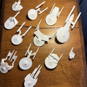 May include: A collection of white, 3D-printed starship models, including various designs with saucer sections and warp nacelles. The models are arranged on a wooden surface, showcasing different sizes and styles of the spacecraft.