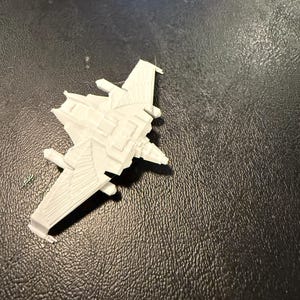 May include: A small, white plastic model of a futuristic aircraft. The model has detailed wings, a central body, and multiple small protrusions. The aircraft is set against a dark, textured background.