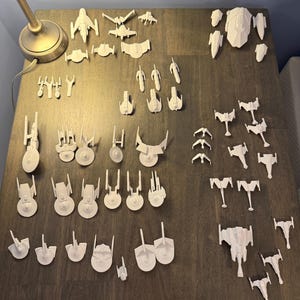May include: A collection of white 3D-printed model spaceships, including various designs of starships and spacecraft. The models are arranged on a dark wooden surface, showcasing different sizes and styles.
