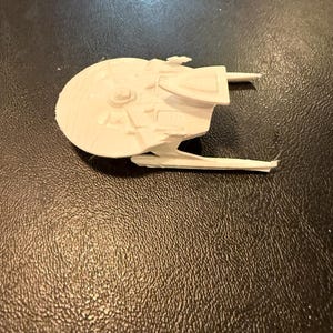 May include: A small, off-white model of a spaceship, featuring a circular main body and extended wings. The model has intricate detailing, including raised panels and a central disc. The spaceship is set against a dark, textured background.
