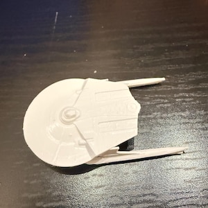 May include: A white, 3D-printed model of a spaceship. The model features a circular main body with two extended wing-like structures. The design includes detailed surface features and a central circular element.