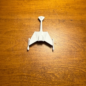 May include: A small, white model of a spacecraft, featuring a central body with wings and a vertical fin. The spacecraft is set against a wooden background. The design suggests a futuristic or science fiction theme.