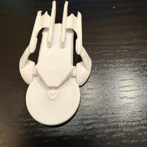 May include: A white 3D-printed model of a spaceship, featuring a circular front section and a rectangular body with multiple vertical fins. The model has detailed structural elements and is set against a dark background.