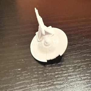 May include: A small, off-white model of a spaceship. The model features a circular base with detailed paneling and a central structure with multiple fins and protrusions. The overall design suggests a futuristic or science fiction theme.