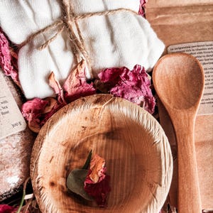 K&ouml;nnte beinhalten: Ein Set bestehend aus einem mit Bindfaden verschn&uuml;rten wei&szlig;en Tuch, einer kleinen Holzschale, einem Holzl&ouml;ffel und getrockneten rosa Rosenbl&auml;ttern. Ein Etikett zeigt die Aufschrift 300 g, ein weiteres beschreibt eine Gesichtsbehandlung in Pulverform.