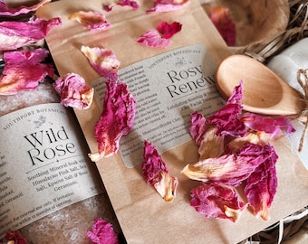 Puede incluir: Una bolsa de papel marrón etiquetada como Rosy Renew que contiene un tratamiento facial y otra bolsa etiquetada como Wild Rose con 300g de sales minerales, rodeadas de pétalos de rosa esparcidos y una pequeña cuchara de madera en una caja.