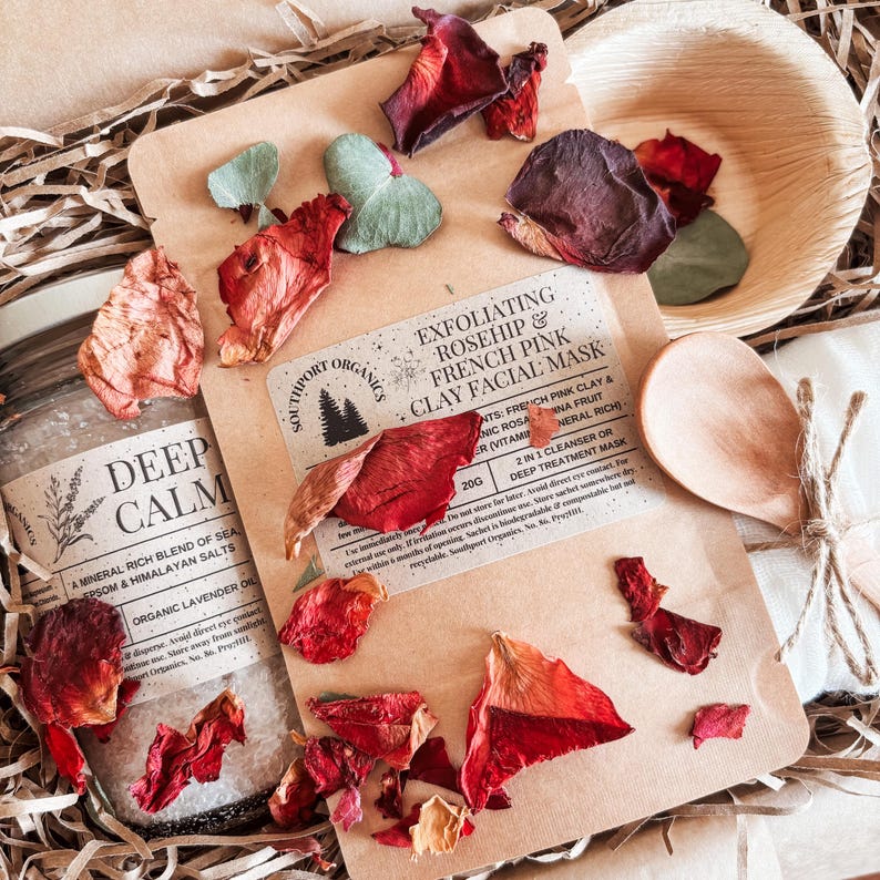 May include: A spa-themed gift set with a jar of Deep Calm bath salts, an exfoliating rosehip and French pink clay facial mask, a wooden spoon, and a small wooden bowl. Dried rose petals and eucalyptus leaves are scattered around the items.