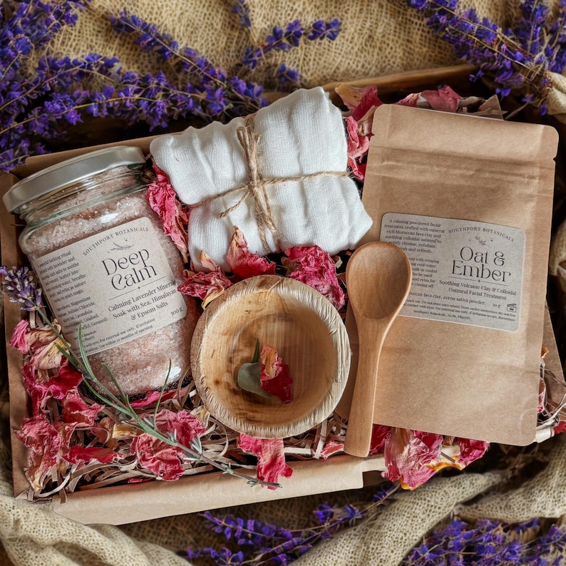 K&ouml;nnte beinhalten: Ein Geschenkset mit einem Glas Deep Calm Lavendel-Badesalz, einem mit Bindfaden verschn&uuml;rten wei&szlig;en Musselintuch, einer kleinen Holzschale und einem Papierbeutel mit Oat & Ember Gesichtsmaske, eingebettet zwischen getrockneten rosa Rosenbl&auml;ttern.