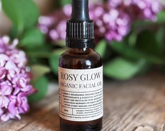 May include: A 30 ml amber glass bottle of Rosy Glow Organic Facial Oil. The bottle has a black dropper top and a white label with the product name and description. The bottle is sitting on a wooden surface with purple flowers in the background.