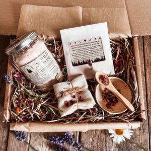 May include: A gift box filled with self-care items. It includes a jar of Wild Rose bath salts, a packet of Rosy Glow exfoliating facial mask, a small wooden spoon, a small wooden bowl, and a wrapped cloth. Dried flowers and lavender are scattered throughout.