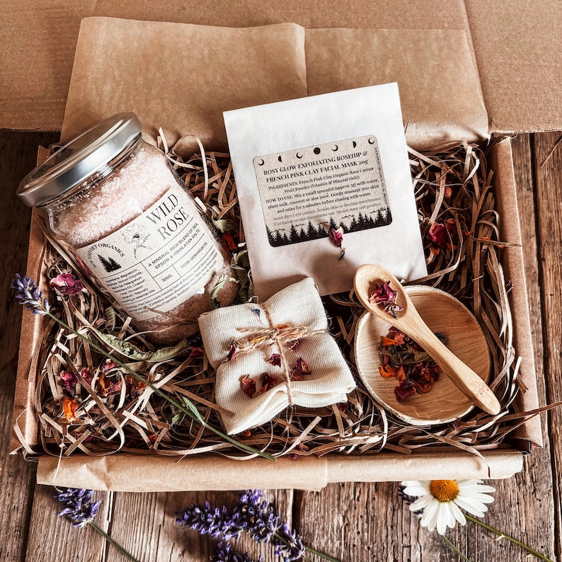 May include: A gift box filled with self-care items. It includes a jar of Wild Rose bath salts, a packet of Rosy Glow exfoliating facial mask, a small wooden spoon, a small wooden bowl, and a wrapped cloth. Dried flowers and lavender are scattered throughout.
