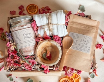 May include: A gift box containing a glass jar of Deep Calm lavender bath soak, a white muslin cloth, a small wooden bowl, and a brown paper pouch of Oat & Ember oatmeal facial treatment, all surrounded by dried flower petals and orange slices.