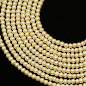 Puede incluir: Primer plano de varios hilos de perlas de imitación de color crema. Las perlas son uniformes en tamaño y forma, con un ligero brillo iridiscente. Los hilos están dispuestos en un patrón curvo sobre un fondo negro, lo que sugiere un collar o una joya similar.