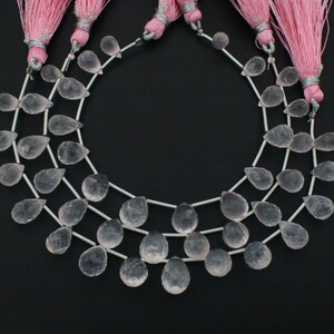 Peut inclure: Trois rangées de perles de pierres précieuses en quartz rose pâle, en forme de goutte d'eau, enfilées sur du fil d'argent. Des pompons roses sont attachés en haut des rangées. Le fond est noir.