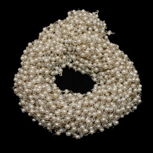 Puede incluir: Un collar de cadena de plata y perlas blancas. La cadena está hecha de pequeños eslabones delicados y está enhebrada con perlas blancas. El collar está enrollado en un círculo.
