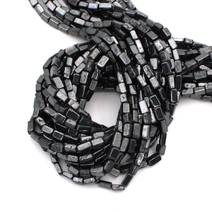 Peut inclure: Gros plan sur des brins de perles d'hématite rectangulaires gris foncé. Les perles ont un éclat métallique et sont enfilées ensemble, créant un aspect texturé. Les perles sont présentées sur fond blanc.