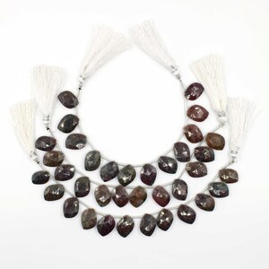 Op de afbeelding: Drie strengen gefacetteerde, donkerroodbruine edelstenen met een peervorm. Elke steen is geregen met een zilverkleurige draad en afgewerkt met een witte kwast. De edelstenen hebben een gepolijst, reflecterend oppervlak.