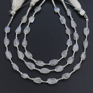 Op de afbeelding: Drie strengen maansteen kralen met een gepolijste, onregelmatige vorm. De kralen zijn geregen op een zilverkleurige draad en afgewerkt met witte kwastjes. De maanstenen hebben een doorschijnende, melkachtige witte kleur met subtiele iriserende effecten.