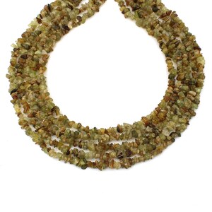 Puede incluir: Una hebra de cuentas de piedras preciosas verdes y marrones, hechas de pequeñas astillas irregulares. Las cuentas están enhebradas en un hilo transparente y están dispuestas en forma circular.