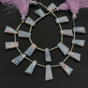 Peut inclure: Un collier de perles en pierre gemme facettées de forme trapézoïdale, bleu clair et rose pâle, alternées avec des tubes cylindriques argentés. Le cordon est terminé par deux pompons en tissu violet clair aux extrémités, formant un bijou.
