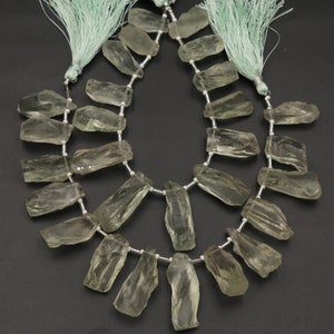 以下が含まれることがあります： 2本の生の淡い緑色の宝石ビーズ。各ストランドは、銀色のワイヤーに連ねられた不規則な形の半透明の石を特徴としています。ストランドは上部に淡い緑色のタッセルで飾られており、自然でアースカラーの美学を作り出しています。