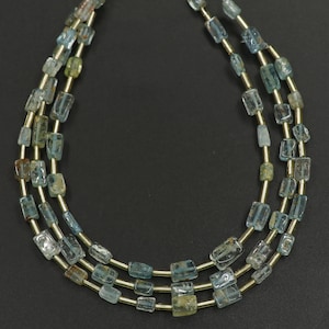 Peut inclure: Un collier à trois rangs orné de perles rectangulaires bleu clair et vertes. Les perles sont enfilées sur un fil doré, créant un effet de superposition. Le collier est présenté sur un fond sombre, mettant en valeur les couleurs et les textures des pierres.