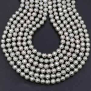 Puede incluir: Primer plano de un collar de cuentas facetadas de color plateado. Las cuentas tienen una superficie texturizada y multifacética, que refleja la luz. Las cuentas están ensartadas, creando un collar o pulsera. El fondo es gris oscuro liso.