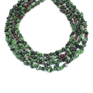 Op de afbeelding: Een ketting gemaakt van meerdere strengen kleine, onregelmatig gevormde groene en kastanjebruine edelstenen. De edelstenen hebben een ruwe, natuurlijke textuur en zijn aan elkaar geregen. De achtergrond is wit.