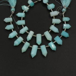 Puede incluir: Una hebra de cuentas de piedras preciosas de color azul claro, cada cuenta está facetada y tiene forma de lágrima larga y estrecha. Las cuentas están enhebradas en un alambre de plata y están separadas por pequeños separadores de plata. Las cuentas están dispuestas en una doble fila, con las cuentas de la fila superior ligeramente desplazadas con respecto a las cuentas de la fila inferior.