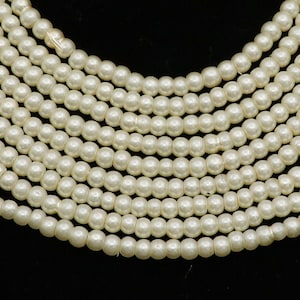 Puede incluir: Primer plano de varios hilos de perlas pequeñas y redondas de color blanquecino. Las perlas están ensartadas, creando un efecto de capas. El fondo es negro liso, resaltando el color y la textura de las perlas.