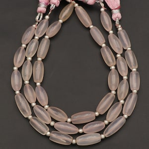 Puede incluir: Tres hilos de cuentas ovaladas de color rosa pálido con espaciadores plateados. Las cuentas están ensartadas juntas, y cada hilo tiene una borla rosa en la parte superior. El fondo es negro liso.