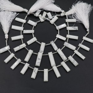 Könnte beinhalten: Stränge aus weißen, rechteckigen Perlen mit grauer Maserung, auf silberfarbenem Draht aufgefädelt. Weiße Quasten sind an den Enden befestigt. Die Perlen sind in einem kreisförmigen Muster vor einem schwarzen Hintergrund angeordnet, für Schmuck.