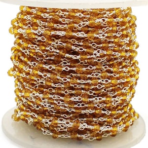Op de afbeelding: Een spoel met kralenketting met kleine, facetgeslepen amberkleurige kralen geregen met zilverkleurig draad. De ketting is strak opgerold en toont het ingewikkelde ontwerp en de kleur.