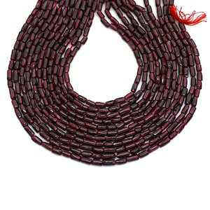 Puede incluir: Hebras de cuentas de piedras preciosas de color rojo burdeos intenso. Las cuentas son cilíndricas con bordes redondeados, ensartadas en un hilo rojo. Las cuentas tienen un aspecto pulido y brillante. El fondo es blanco.