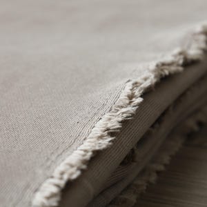 Puede incluir: Primer plano de una pila de tela beige con un borde deshilachado. La tela es suave y texturizada, con un brillo sutil.
