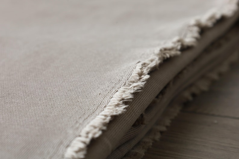 Puede incluir: Primer plano de una pila de tela beige con un borde deshilachado. La tela es suave y texturizada, con un brillo sutil.