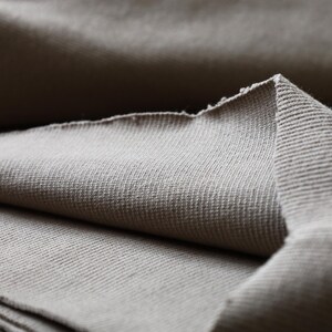 Puede incluir: Primer plano de una tela suave de color beige con textura acanalada. La tela est&aacute; doblada por la mitad, creando una superficie lisa y uniforme.