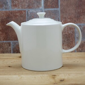 May include: A white ceramic teapot with a lid and handle, set against a brick wall background. The teapot has a spout for pouring and a small knob on the lid. The teapot is sitting on a wooden surface.